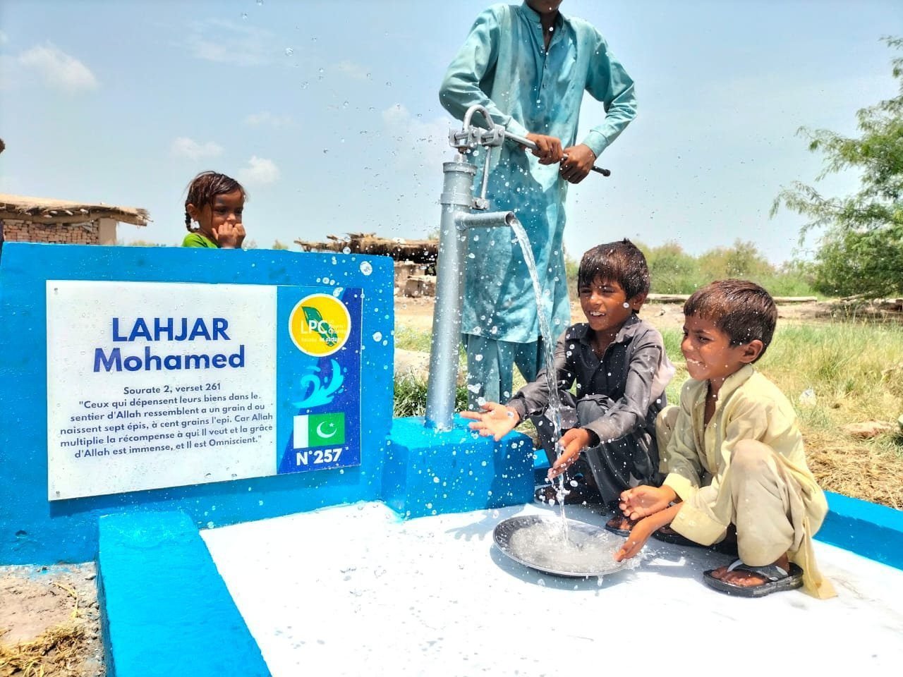 Children drinking at a blue hand-pump well in Pakistan, named plaque in front