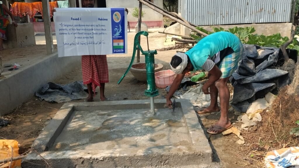 Worker finishing the concrete platform of a hand-pump well, with a plaque carrying a dua for the customer's parents