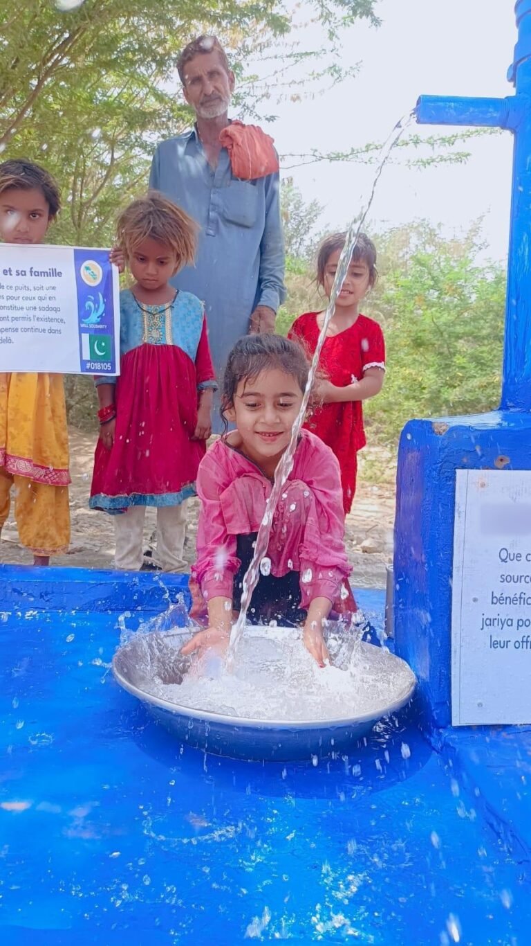 Children splashing at a new hand-pump water well in Sindh, Pakistan, built by Well & Trees, with the named plaque