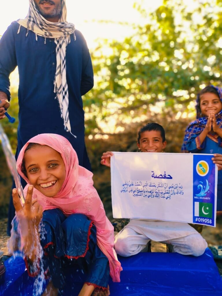 Smiling girl at a hand-pump well in Pakistan, boy holding a plaque written in Arabic