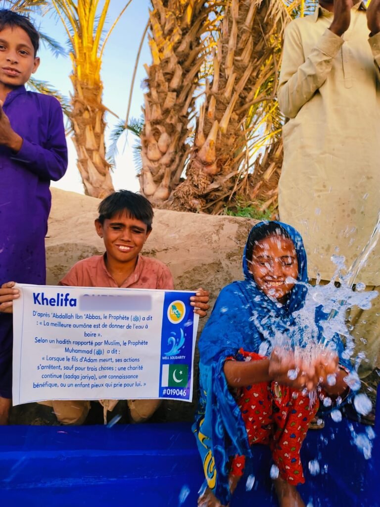 Boy holding a plaque with the hadith of the three deeds at a hand-pump well in Pakistan, girl splashing water