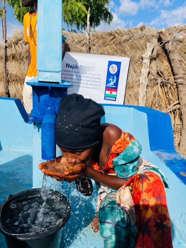 Child drinking from a hand-pump well in Niger, plaque in memory of a deceased person