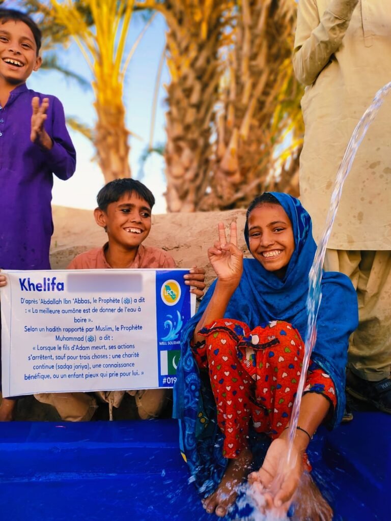 Two children smiling at a hand-pump well in Pakistan, holding the customer's plaque, water splashing