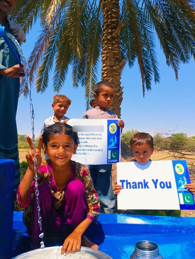 Children at a water well in Pakistan holding the named plaque and a Thank You sign under a palm tree