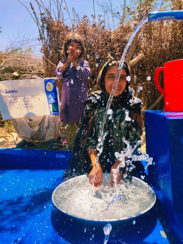 Girl filling a basin at a hand-pump well in Pakistan, red jug on the well head, plaque held behind