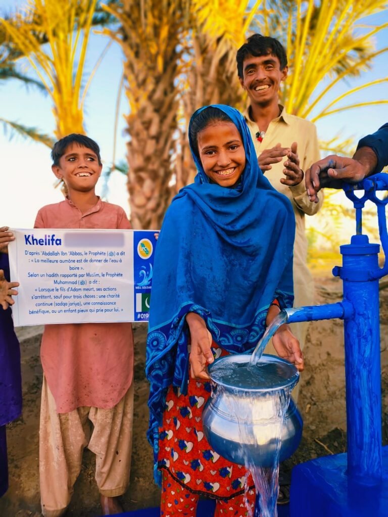 Girl in a blue scarf filling a metal pot at a blue hand-pump well in Pakistan, boy holding the plaque
