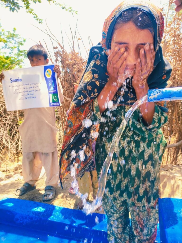 Girl washing her face in the stream of a blue hand-pump well in Pakistan, boy holding a plaque with a hadith