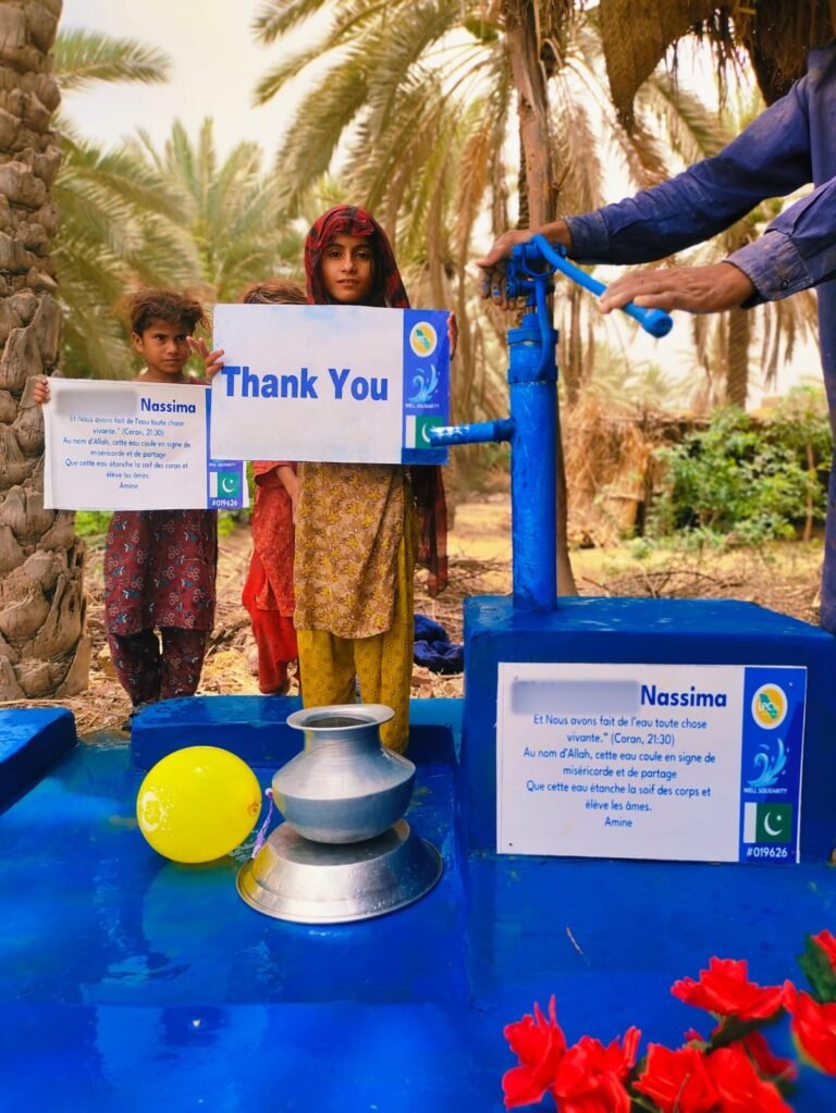 Hand-pump well in Pakistan with a plaque quoting Qur'an 21:30, children holding a Thank You sign, flowers and a balloon